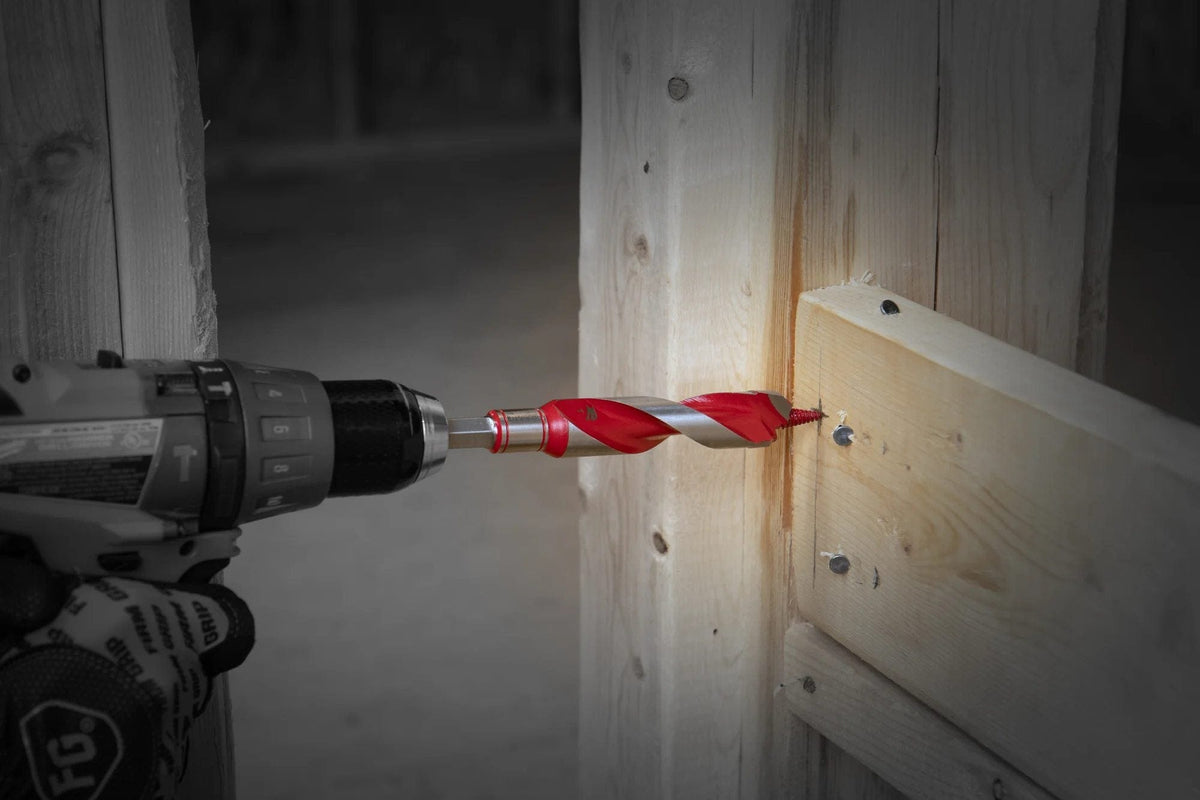 An action photograph of a handheld power drill equipped with a red and silver 3/4 inch Diablo auger bit boring through a thick structural timber and successfully cutting through an embedded metal nail.