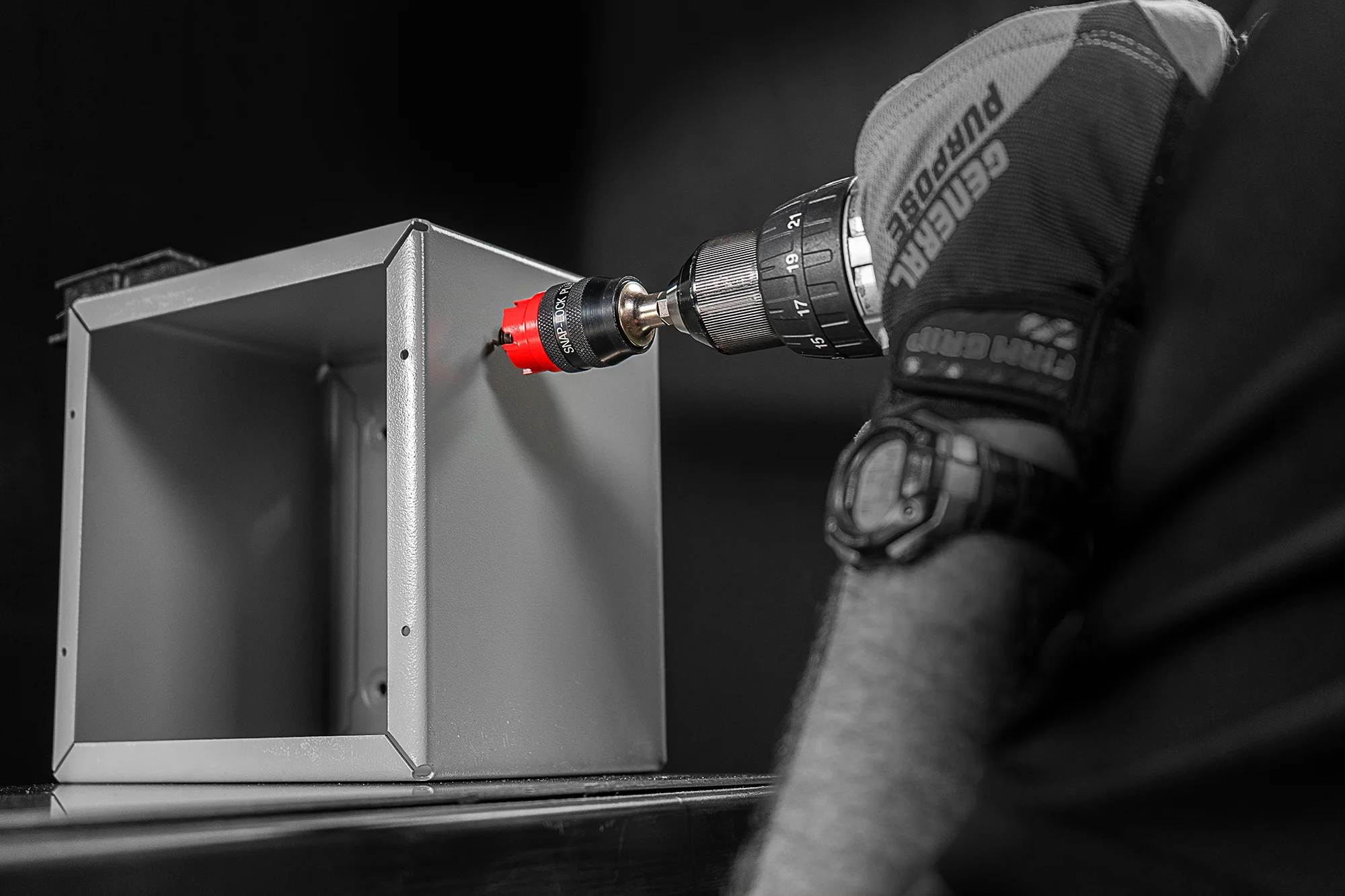 A professional action shot in greyscale with selective red coloring. An electrician in a safety glove uses a handheld drill equipped with a red Diablo carbide hole cutter to bore into a grey metal electrical junction box.