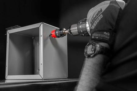 A professional action shot in greyscale with selective red coloring. An electrician in a safety glove uses a handheld drill equipped with a red Diablo carbide hole cutter to bore into a grey metal electrical junction box.