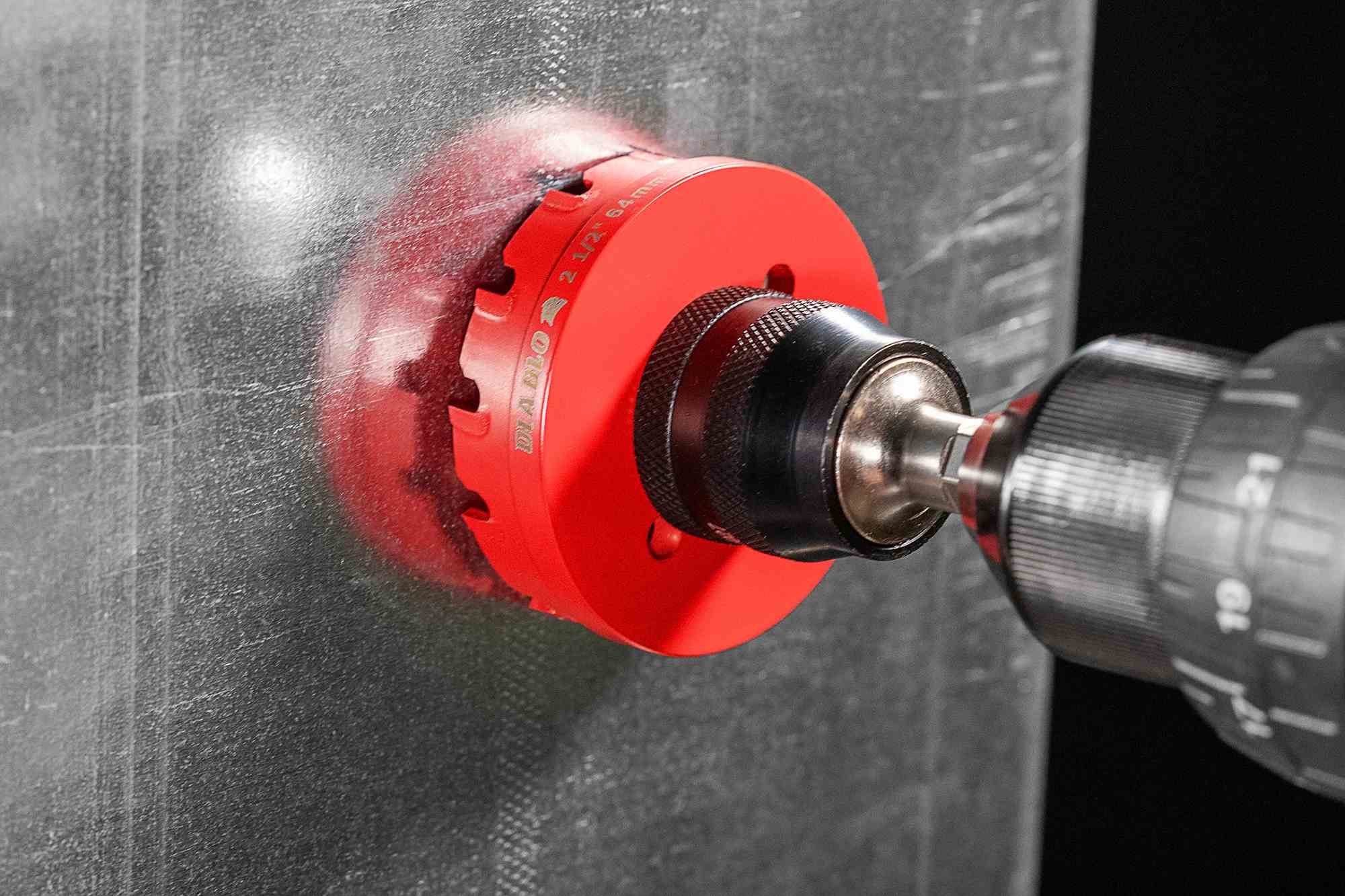 A close-up action photograph of a red Diablo Steel Demon hole cutter boring through a flat sheet of stainless steel. The image highlights the clean, burr-free entry point and the tool's efficiency in metal fabrication.