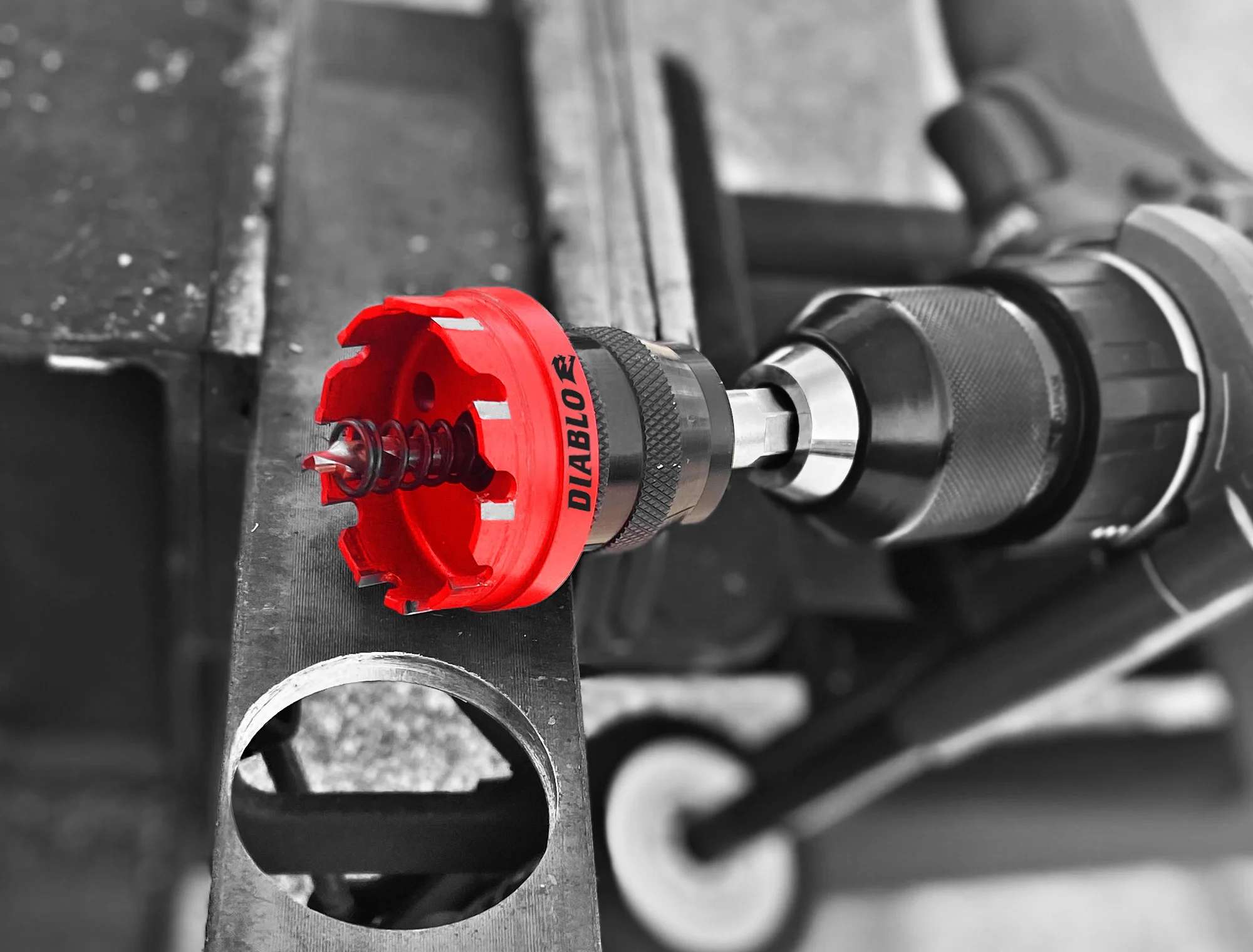A greyscale action shot with selective red coloring showing a Diablo carbide hole cutter attached to a handheld drill. The cutter is being used to bore through a thick metal structural beam, demonstrating high-performance metal removal.