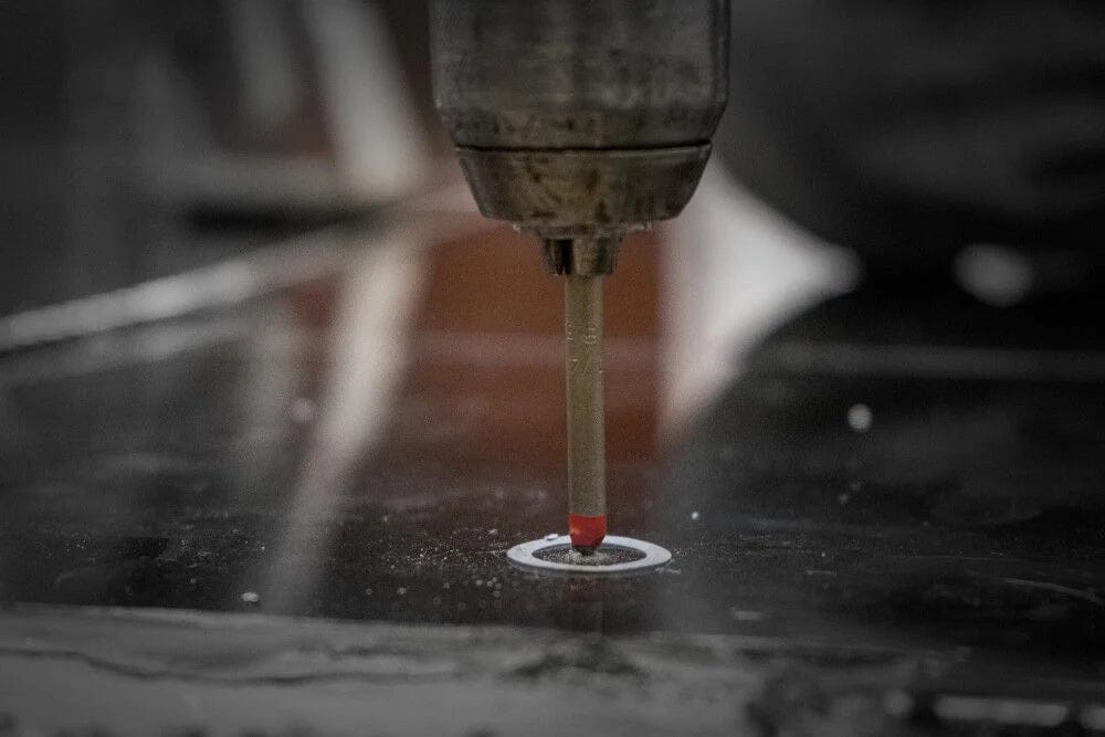 An action shot showing a specialized drill bit with a red multi-ground carbide tip boring a clean hole into a dark, smooth glass or ceramic surface.