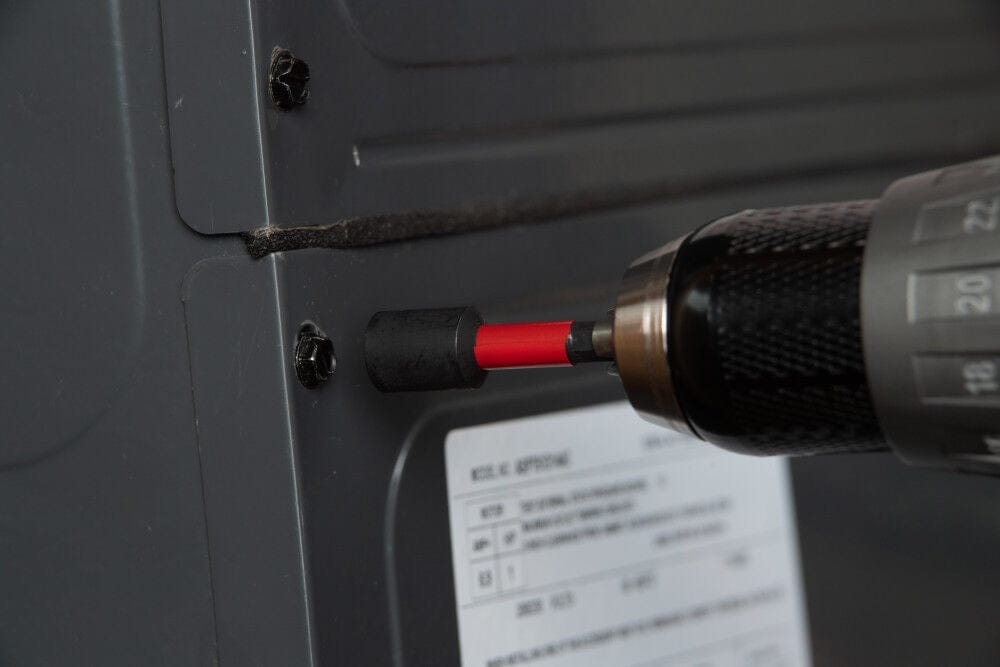 An action shot of a hand-held cordless drill using a Diablo magnetic nut setter to drive a black hex-head screw into a gray metal panel. The nut setter features a prominent red band between the black socket head and the drill chuck.