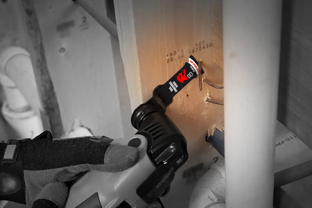 An on-site application photograph of a worker wearing protective gloves using an oscillating multi-tool fitted with the black and red Diablo bi-metal blade to make a precise flush cut through a wood framing stud embedded with thick steel construction nails.