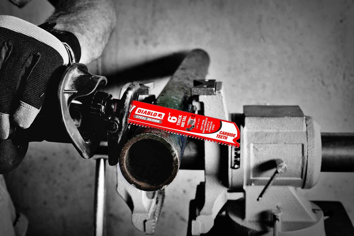 An action photograph of a 6 inch Diablo Steel Demon carbide blade in a reciprocating saw cutting through a thick-walled metal pipe held in a workbench vice, demonstrating the blade's industrial performance and high-heat resistance.