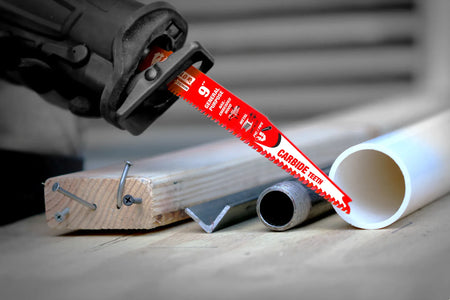 An in-use action photograph of a reciprocating saw running the red Diablo carbide blade down into an assortment of demolition materials, including nail-embedded construction lumber, iron angle stock, steel conduit pipe, and white plastic PVC plumbing.