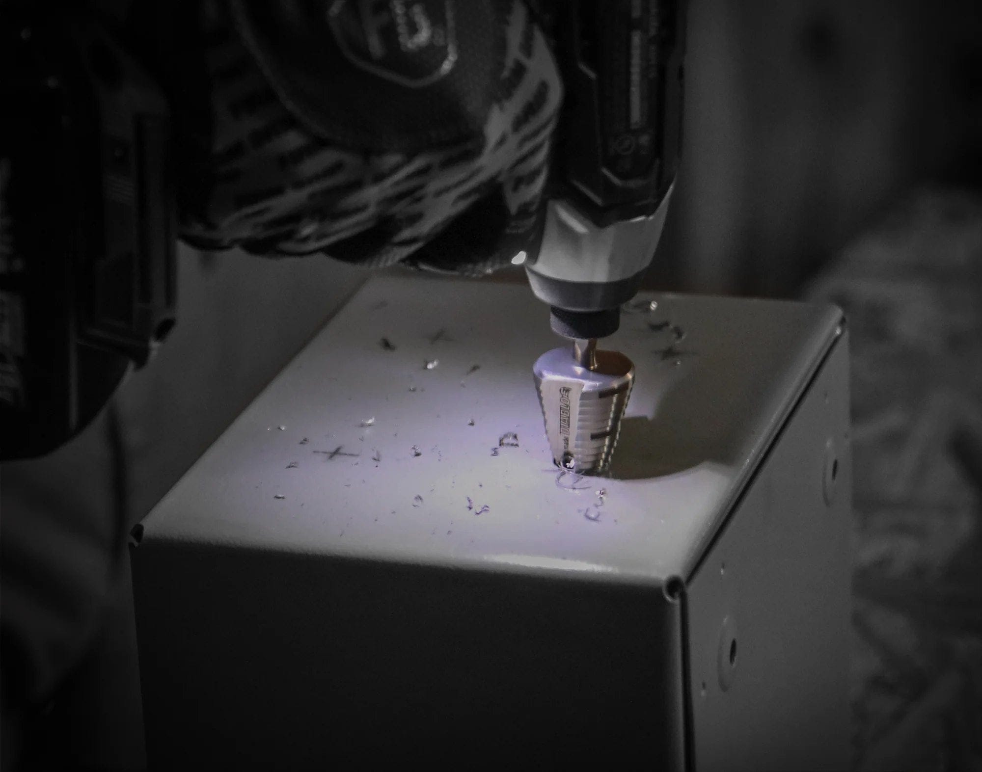 Action shot of a cordless impact driver using the Diablo step bit to drill a hole into a gray metal electrical junction box, with metal shavings visible around the cut.