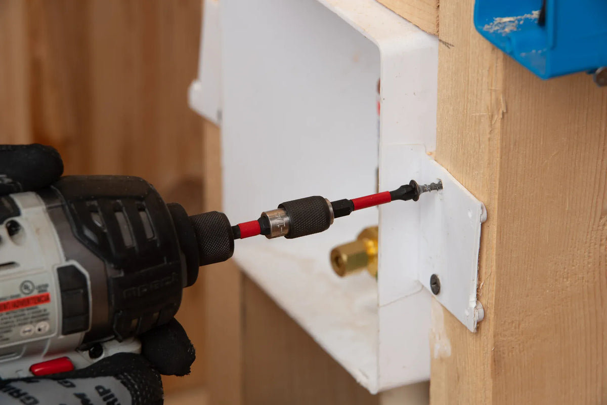 An action close-up shot of a professional tradesperson using a cordless impact driver fitted with an extended magnetic bit holder and a Diablo Phillips driver bit. The tool is driving a fastener through the structural mounting flange of a white utility box directly into a wooden framing stud on a residential construction job site.