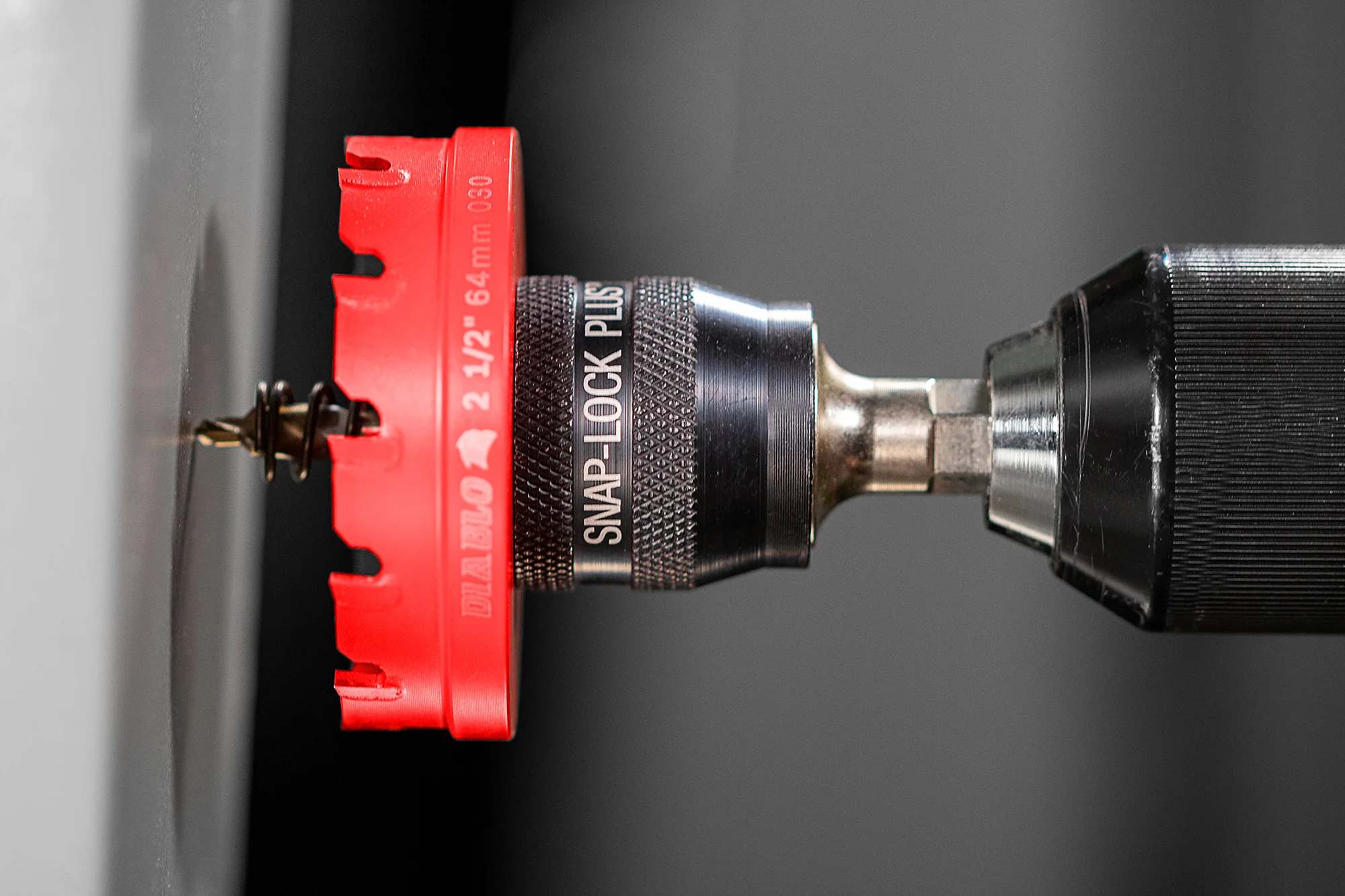 A close-up action shot of a red 2 1/2-inch Diablo hole saw with a Snap-Lock Plus mandrel drilling a hole into a gray metal surface.