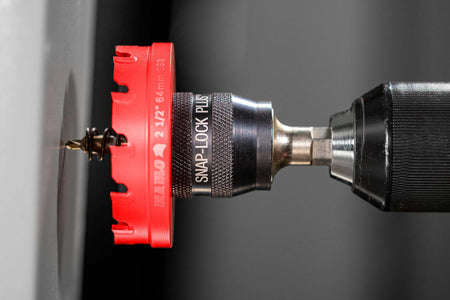 A close-up action shot of a red 2 1/2-inch Diablo hole saw with a Snap-Lock Plus mandrel drilling a hole into a gray metal surface.