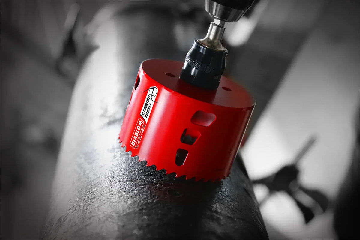 A red Diablo carbide hole saw mounted on a drill tool boring a hole into a dark metal pipe showing the cutting teeth in contact with the material