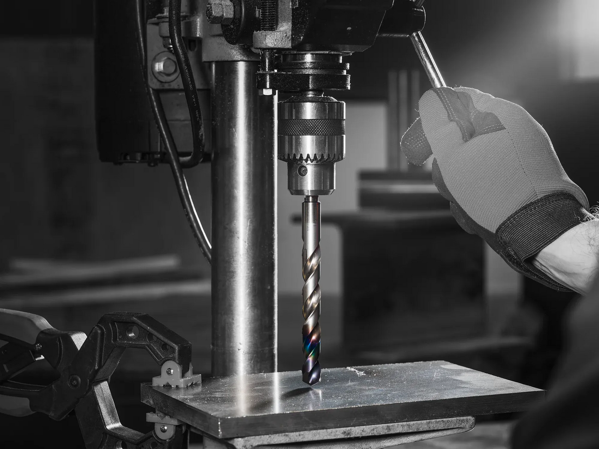 A hand in a work glove operating a drill press with a Diablo Metal Demon drill bit creating a hole in a metal sheet secured with a clamp