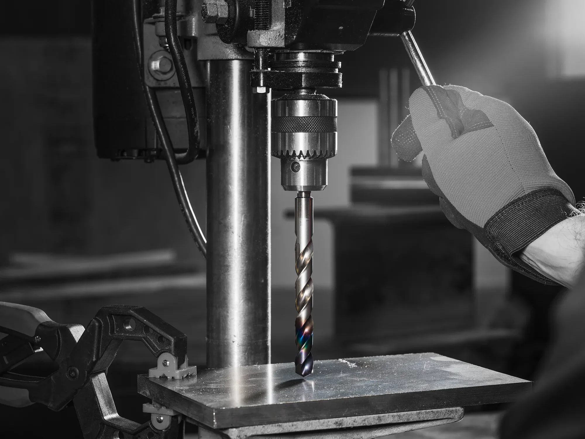 Hand in work glove operating a drill press with a Diablo Metal Demon drill bit creating a hole in a metal sheet secured with a clamp