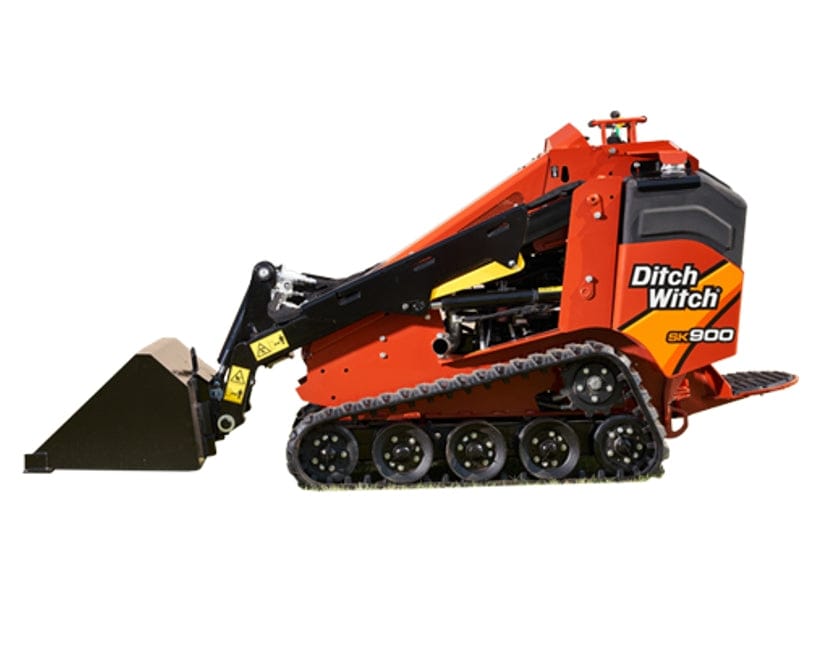 Compact Ditch Witch SK900 mini skid steer loader parked on a job site, featuring rubber tracks, front-mounted bucket, and hydraulic arms; designed for landscaping, construction, and tree care tasks. Branding is visible on the side of the machine.