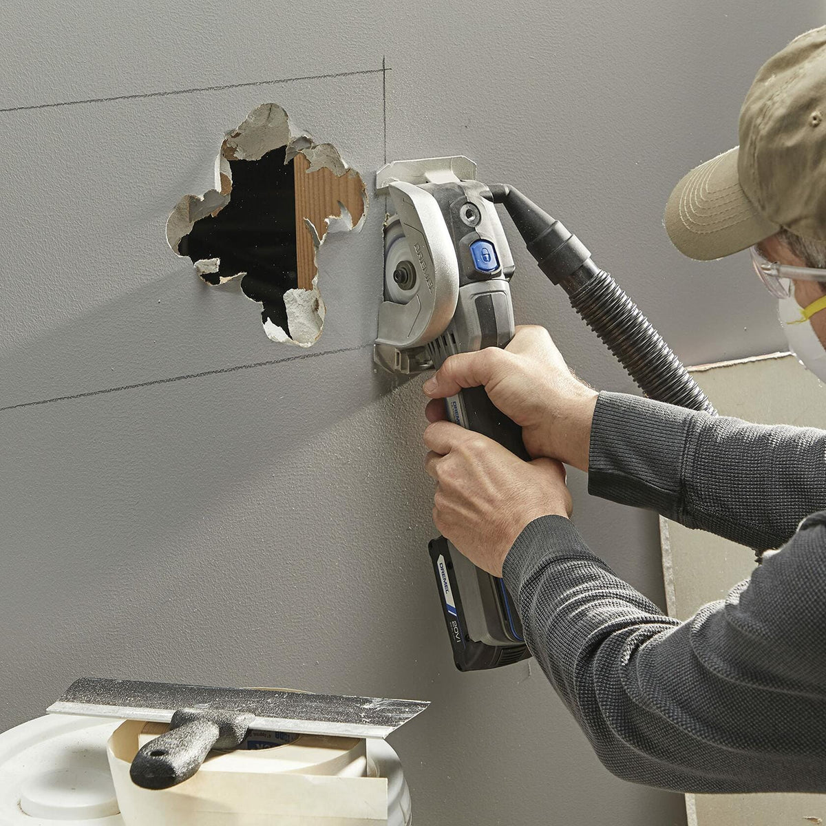 A person making a plunge cut into a gray drywall wall with the Dremel Ultra-Saw, which has a dust collection hose attached for a cleaner workspace.