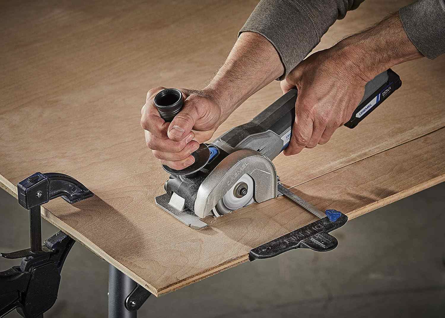 The Dremel US20V compact saw in action, with a user guiding it along a straight edge to cut a piece of clamped plywood.