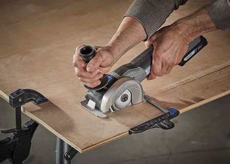 The Dremel US20V compact saw in action, with a user guiding it along a straight edge to cut a piece of clamped plywood.