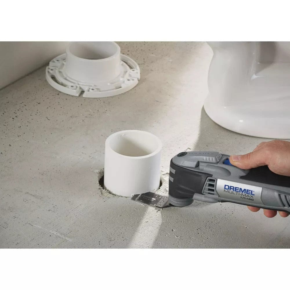 A hand holds the Dremel Multi-Max MM40, using a flush-cut blade to cut a white PVC pipe level with a concrete floor.