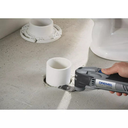 A hand holds the Dremel Multi-Max MM40, using a flush-cut blade to cut a white PVC pipe level with a concrete floor.