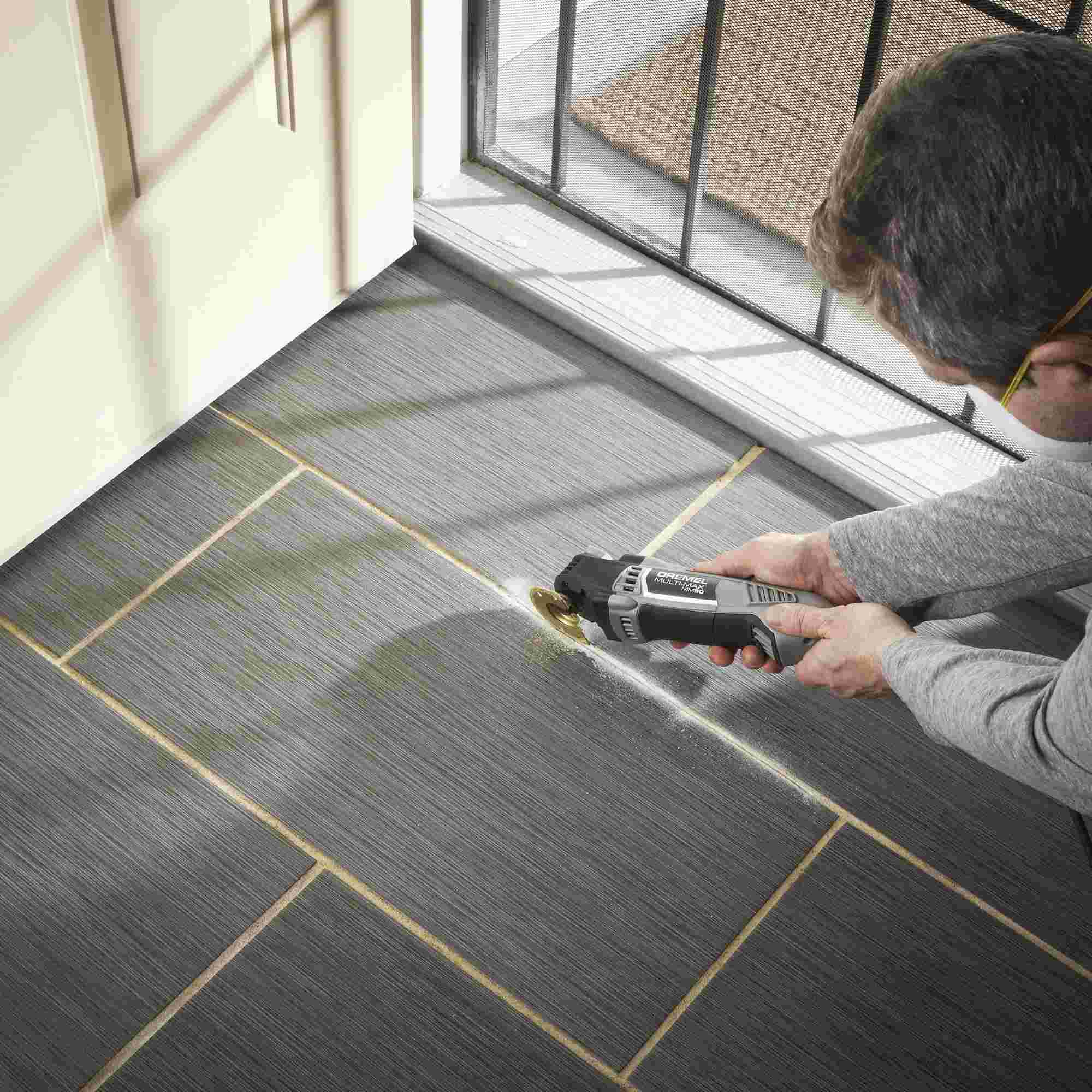 A person uses the Dremel Multi-Max MM50 with a half-moon grout removal blade to remove old grout from between gray floor tiles.