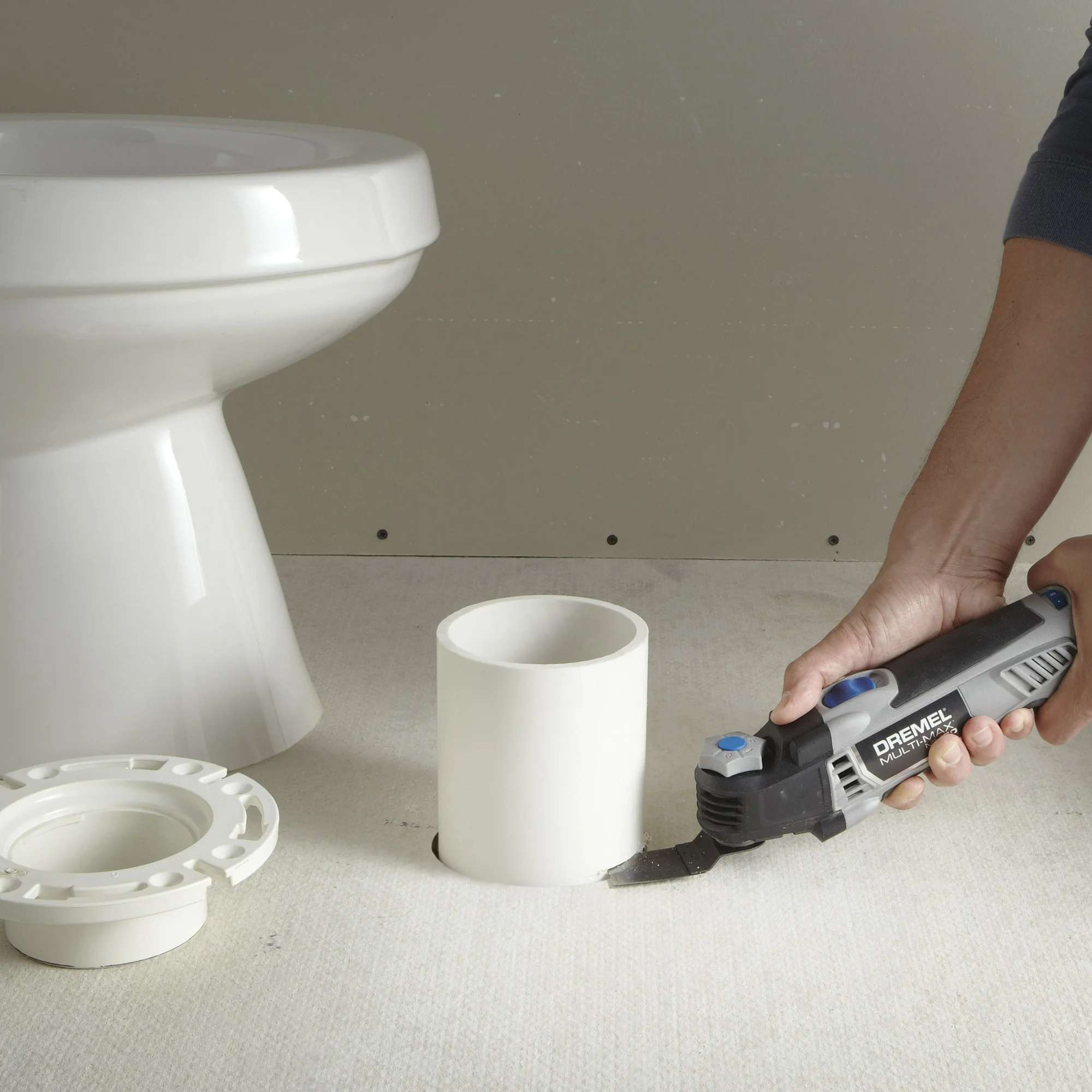 A person uses the Dremel Multi-Max MM50 with a flush cut blade to cut a PVC pipe level with the floor, next to a toilet flange.