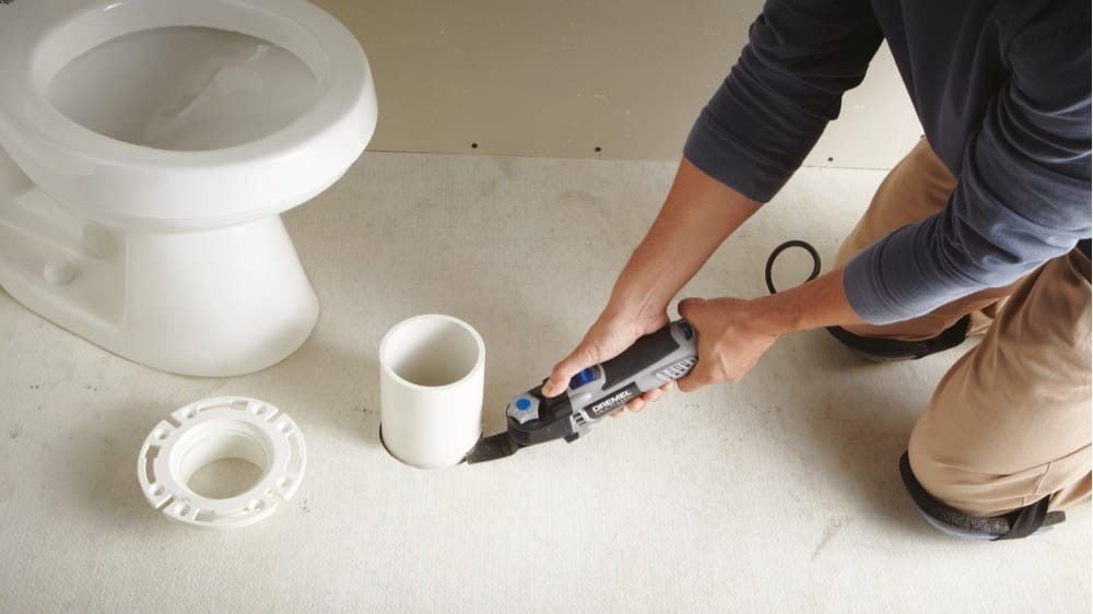 Action shot of a professional plumber using the Dremel Multi-Max MM50 to perform a precise flush cut on a closet flange during a bathroom flooring installation.
