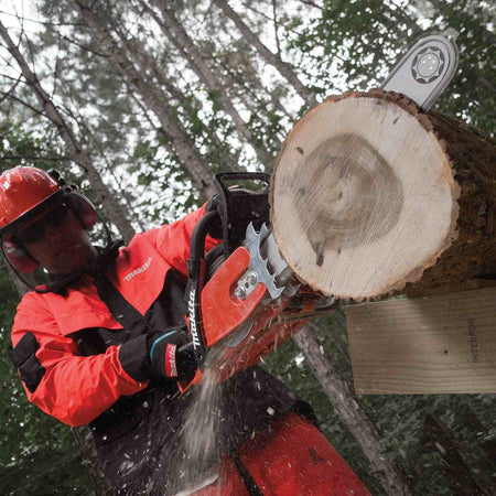 A user wearing orange protective safety gear cuts through a large fallen log with the Makita chainsaw, creating a spray of sawdust.