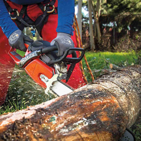 A user wearing orange protective safety gear cuts through a large fallen log with the Makita chainsaw, creating a spray of sawdust.