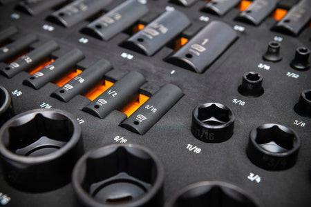A foam tray with a wooden base holds a variety of impact sockets, extension bars, and accessories. Sockets range from 3/8" to 1-1/2" and are arranged in rows by size and depth. Designed for organized access in professional mechanical work.