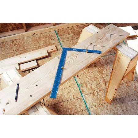 An overhead jobsite application lifestyle photo of the blue Empire framing square resting across a raw dimensional lumber board on wooden sawhorses, with brass stair gauges clamped onto the blade edges to mark out a repetitive stair stringer layout.