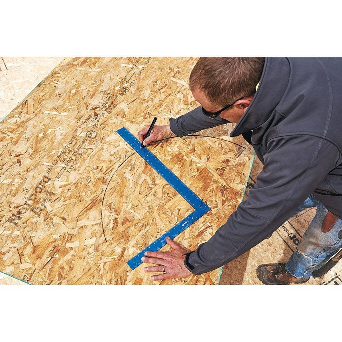 An overhead action application photo of a carpenter using his left hand to hold the blue anodized layout square flat against a large sheet of textured structural strand board while his right hand uses a construction pencil to scribe a uniform curved arc line.
