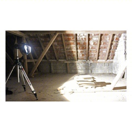 Tripod-mounted Bosch LED floodlight illuminating a dusty attic or unfinished room with red brick ceiling and exposed wooden beams. Light casts shadows across stacked boards and concrete block walls. Highlights jobsite lighting performance in construction or renovation environments.