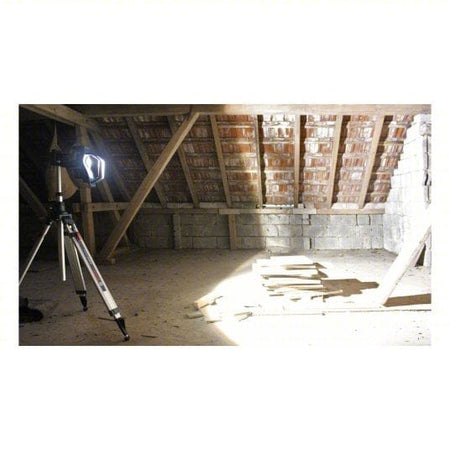 Tripod-mounted Bosch LED floodlight illuminating a dusty attic or unfinished room with red brick ceiling and exposed wooden beams. Light casts shadows across stacked boards and concrete block walls. Highlights jobsite lighting performance in construction or renovation environments.