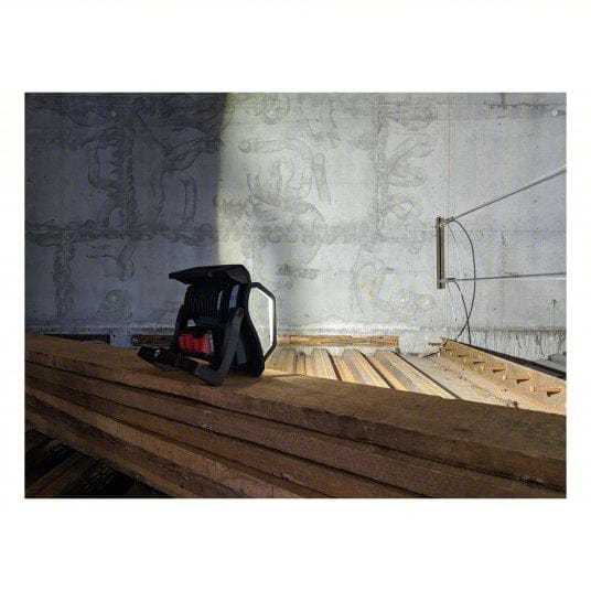 Tripod-mounted Bosch LED floodlight illuminating a dusty attic or unfinished room with red brick ceiling and exposed wooden beams. Light casts shadows across stacked boards and concrete block walls. Highlights jobsite lighting performance in construction or renovation environments.