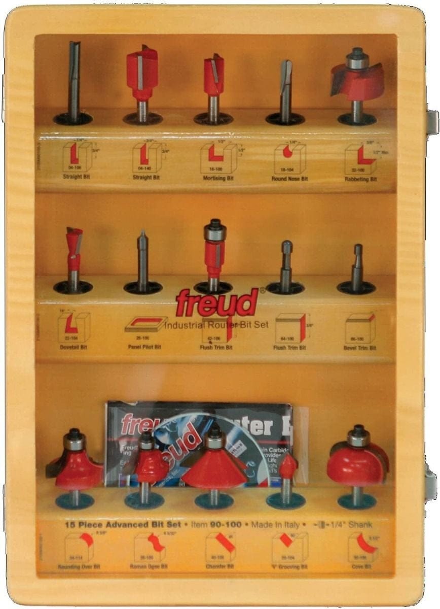 Freud 15-piece industrial router bit set displayed in an open wooden case. Bits are vertically mounted and labeled with type and function, including straight, mortising, flush trim, Roman ogee, rounding over, and dovetail profiles