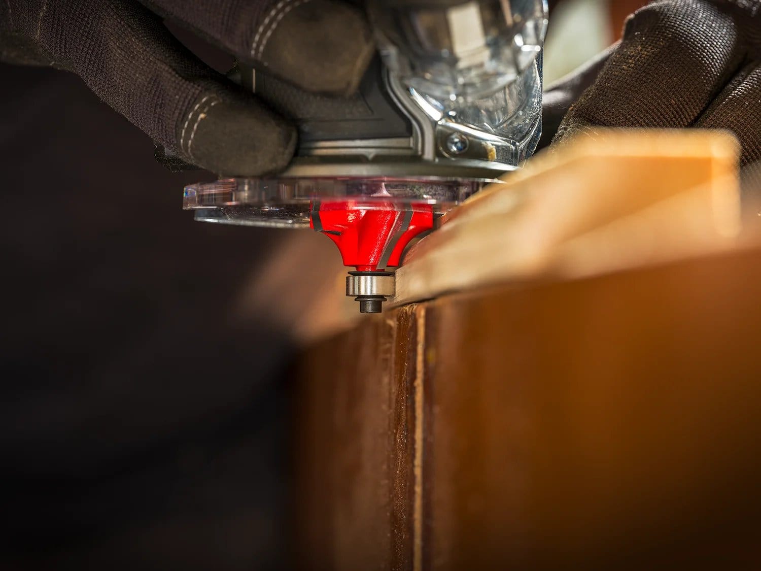 A close-up of a worker wearing gloves using a handheld router with the red Freud beading bit to rout a decorative edge on a wooden workpiece.