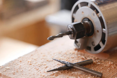 Power drill with attached bit on wood surface, surrounded by wood shavings and two additional carving bits.