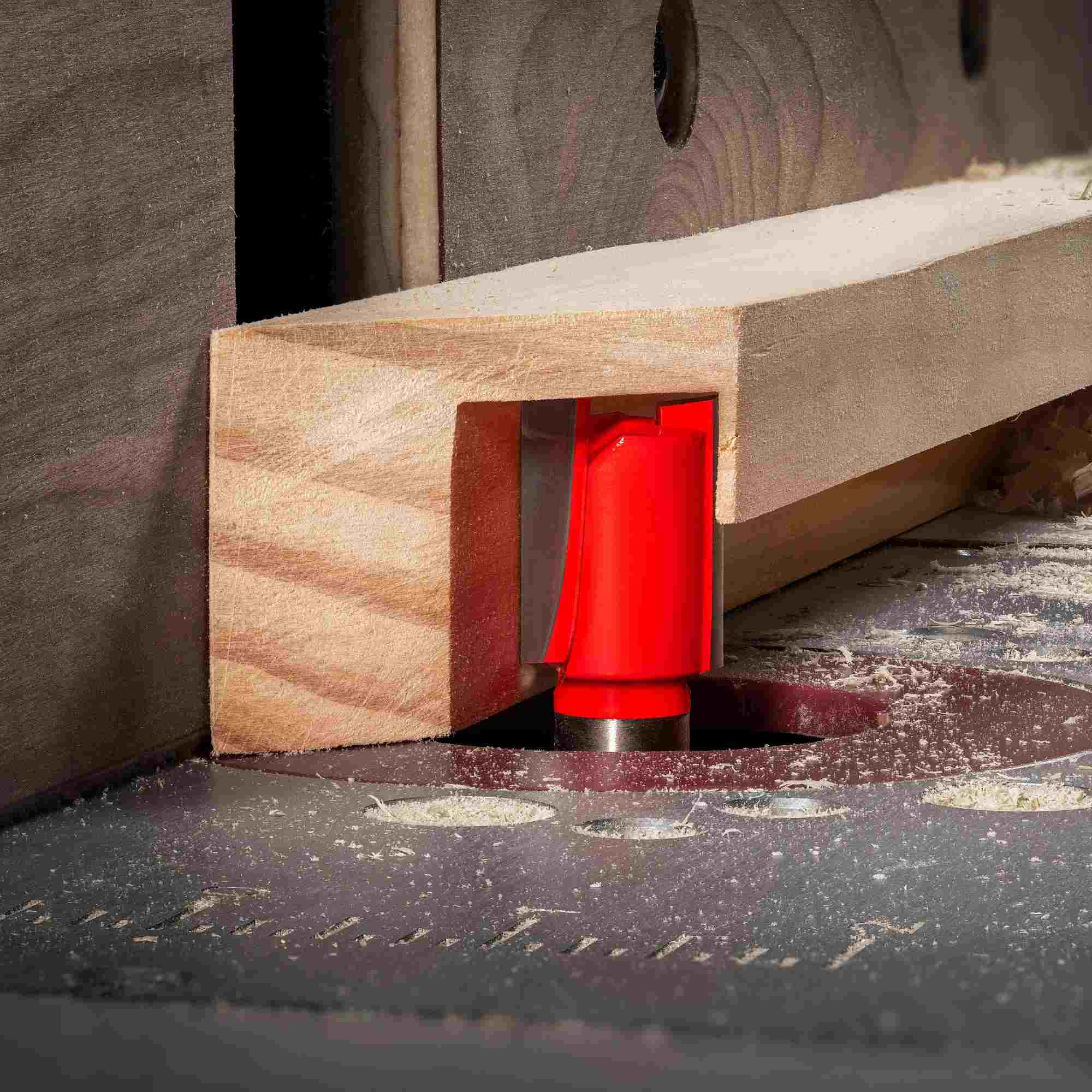 Action shot of the Freud 12-136 router bit with red Perma-SHIELD coating installed in a table router, actively cutting a precise 5/8 inch wide dado groove into a light wood block, with wood shavings and sawdust visible on the grey router table surface and wooden fence.
