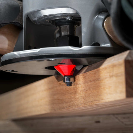 An action-oriented close-up of a Freud chamfer bit mounted in a plunge router, demonstrating the tool creating a crisp 25-degree bevel edge on a hardwood workpiece.