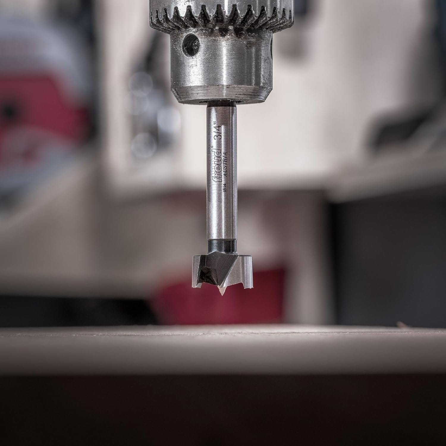 A close-up action shot of a Freud precision shear Forstner bit mounted in a drill press chuck, emphasizing the ultra-sharp center spur and serrated edges positioned above a workpiece.