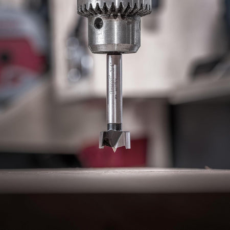 A close-up action shot of a Freud precision shear Forstner bit mounted in a drill press chuck, emphasizing the ultra-sharp center spur and serrated edges positioned above a workpiece.