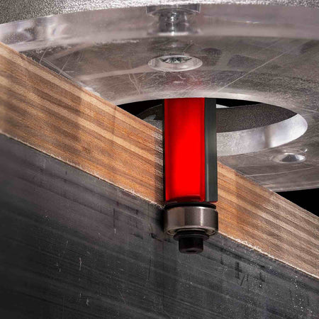 Close-up action shot of the Freud 50-509 bearing flush trim bit mounted in a router, actively trimming a plywood edge perfectly flush with the bottom ball bearing pilot.