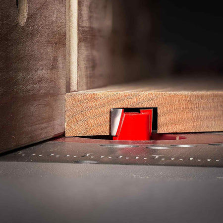 Close-up action shot of a red Freud router bit cutting a clean, precise dado groove into a light wood board during a router table operation.