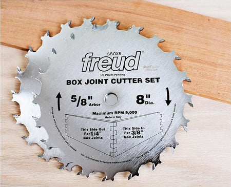 Lifestyle image of the Freud 8-inch box joint cutter blades resting on a piece of hardwood, demonstrating the professional industrial design and rotation direction for woodworking.