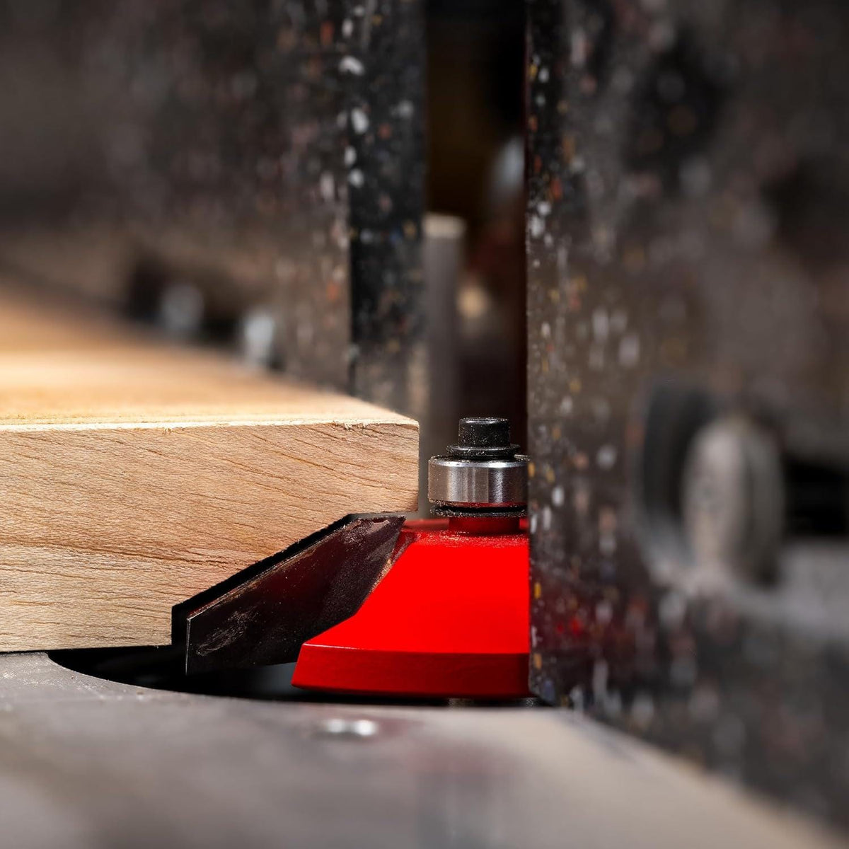 Close-up action shot of a table-mounted router equipped with the red Freud Quadra-Cut raised panel router bit shaping a smooth, tear-out-free profile on a solid wood panel.