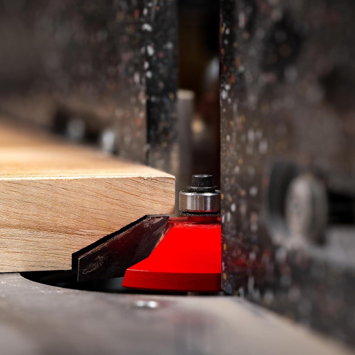 Close-up action shot of a table-mounted router equipped with the red Freud Quadra-Cut raised panel router bit shaping a smooth, tear-out-free profile on a solid wood panel.