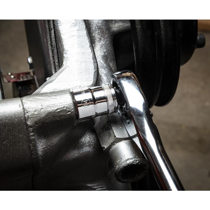 A ratchet wrench engaged with a bolt on a metallic engine component. The socket fits securely over the bolt head, illustrating practical use of the GEARWRENCH socket set in mechanical or automotive repair.