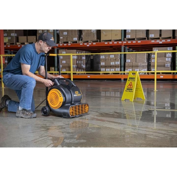 A person kneels on a wet warehouse floor using a black and orange GEARWRENCH air mover to dry the surface. A yellow “Caution Wet Floor” sign is placed nearby for safety.