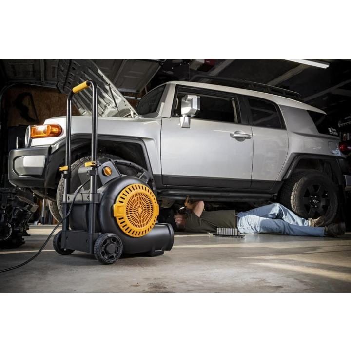 A person works beneath a Toyota FJ Cruiser in a garage, with a yellow and black GEARWRENCH air mover positioned nearby. The fan is plugged in and ready for use, highlighting its role in automotive maintenance.