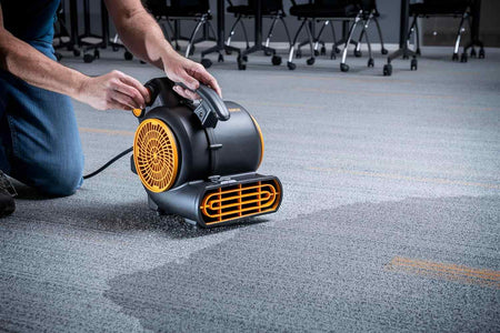 Alt Text: A GEARWRENCH air mover is positioned on a carpeted floor, actively drying a visibly wet patch. Office chairs are visible in the background, suggesting use in a commercial space.
